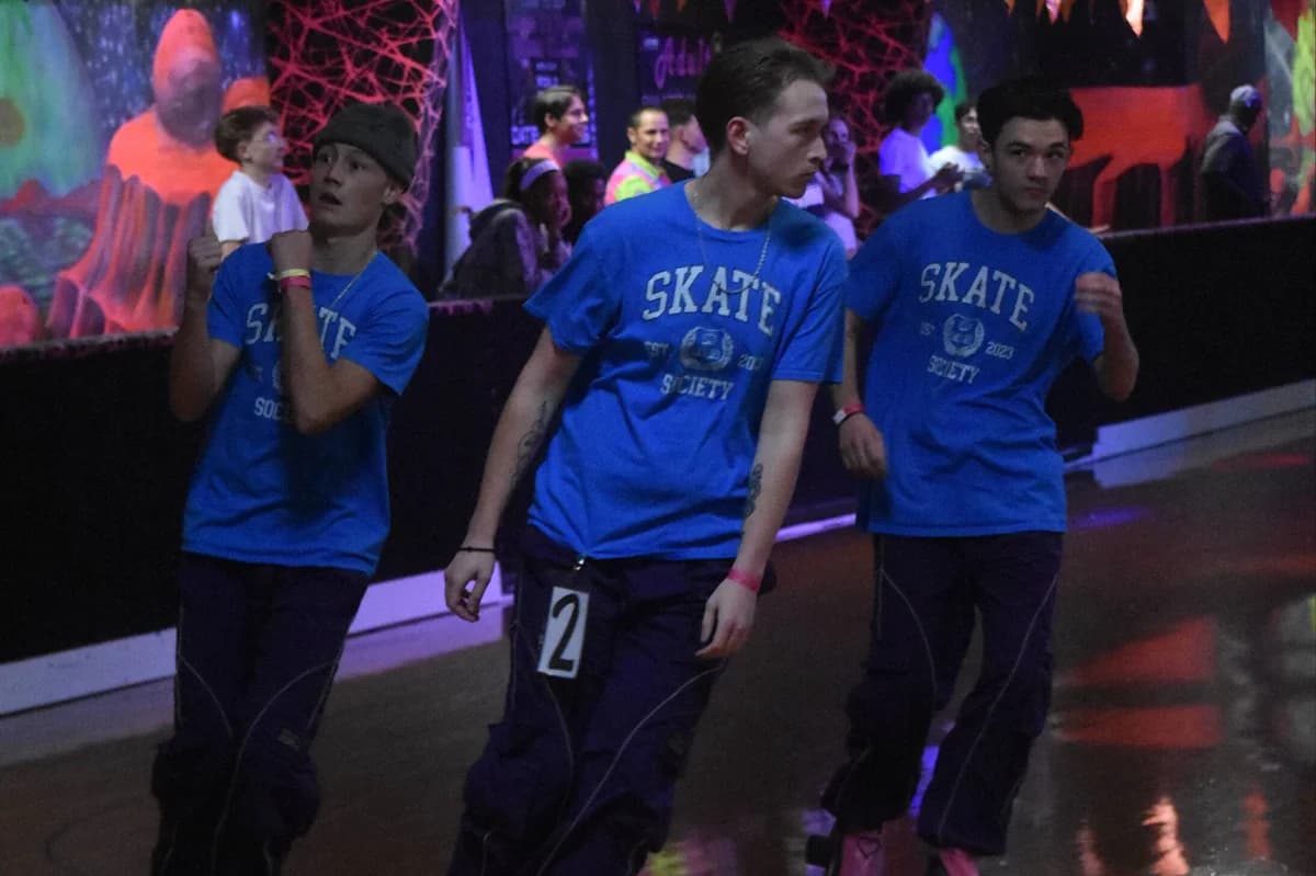 Three Skate Society skaters in matching blue crew tees line up on the rink floor.